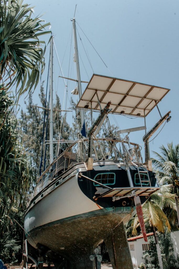 A sailboat being repaired on land surrounded by tropical trees, under a clear blue sky.